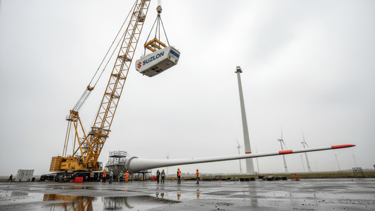 A large yellow crane lifts a white Suzlon wind turbine nacelle above workers and a long white turbine blade on a wet, grey tarmac.