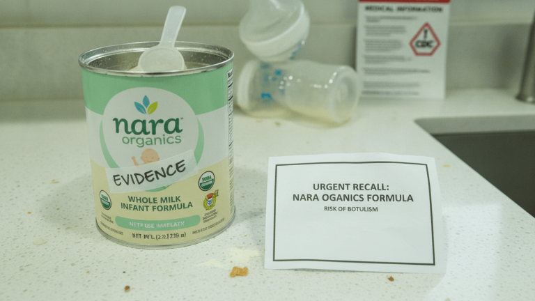 An open can of Nara Organics infant formula with a scoop, labeled "EVIDENCE," sits on a counter next to an urgent recall notice.