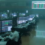 A wide shot of a large trading floor with multiple traders sitting at desks, each with several computer screens displaying financial data. A large screen at the back shows "CRISIL LIMITED SALES ALERTS UPDATES."