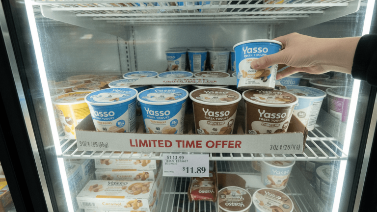 A person's hand reaches into a brightly lit Costco freezer, selecting a single cup of Yasso Frozen Greek Yogurt. Rows of 12-packs of yogurt cups are visible on shelves, with price signage.