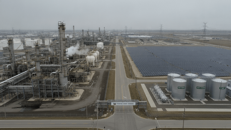 An aerial view of a large industrial complex featuring an oil refinery with storage tanks and a sprawling solar panel array under a dull, overcast sky.