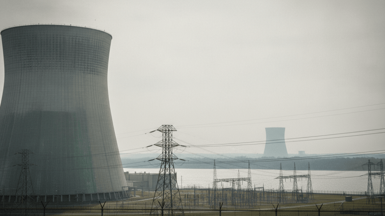 A wide shot of a nuclear power plant with two cooling towers, power lines, and a body of water under an overcast sky.
