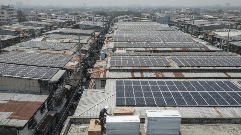 An aerial view of a dense urban area in Manila shows countless corrugated metal rooftops, many fitted with solar panels. In the foreground, a worker wearing a hard hat unboxes new solar panels on a partially installed roof.