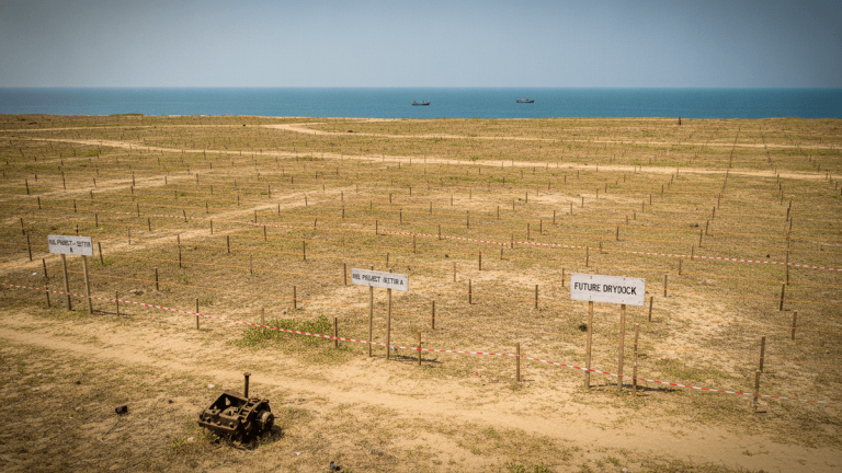 Wide shot of a large, arid coastal plot marked with white signs indicating "FUTURE DRY DOCK" and "HSL PROJECT - SECTOR A". Two small boats are visible on the distant horizon of the blue sea. A rusted piece of machinery sits in the foreground.