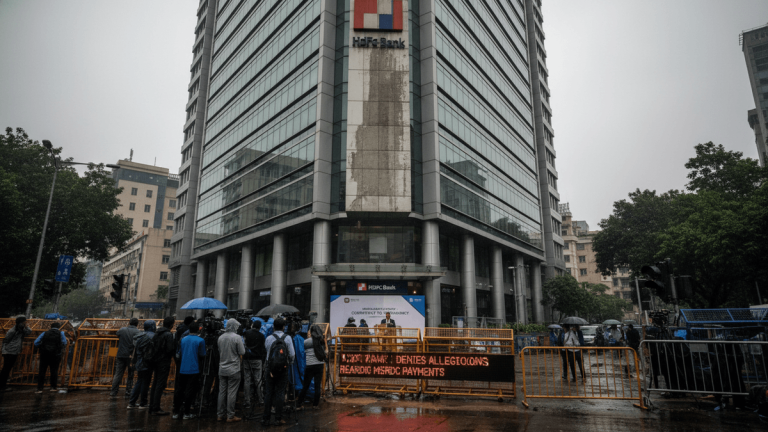 A wide shot of the HDFC Bank building with reporters and photographers gathered in front, a news ticker displaying "HDFC BANK DENIES ALLEGATIONS REGARDING MSRDC PAYMENTS" is in the foreground.