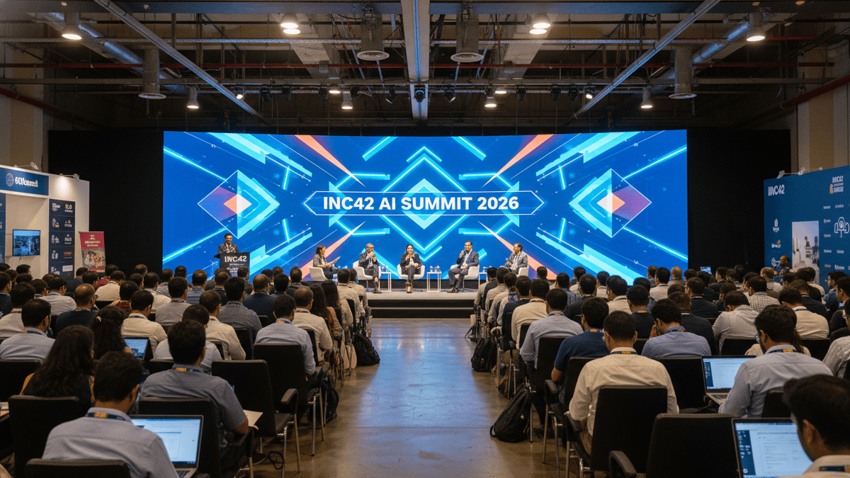 Wide shot of a large conference hall with a panel on stage and a seated audience in front of a brightly lit screen.