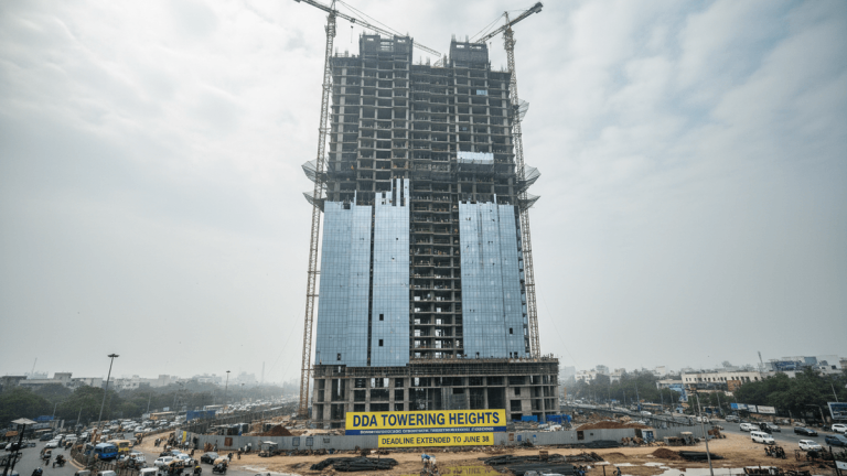 A towering, unfinished skyscraper dominates the frame, surrounded by urban sprawl and busy street traffic under an overcast sky.