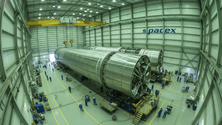 An ultra-wide shot of a massive industrial building with the SpaceX logo. Two large, cylindrical rocket booster sections dominate the foreground, with numerous technicians in blue jumpsuits working around them.
