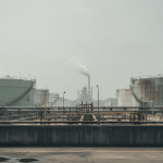 A wide shot of a large oil refinery with multiple white and light green storage tanks, interconnected by a network of pipes.