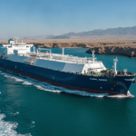 A blue and white LNG tanker moves through a strait with arid coastlines and mountains under a clear sky.