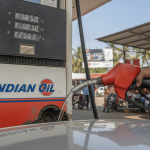 A person's hand holds a red fuel pump nozzle dispensing petrol into a vehicle, with the Indian Oil pump display showing prices.