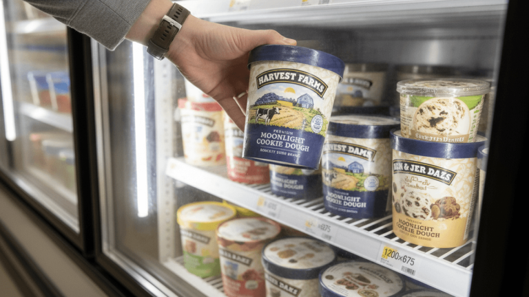 A hand reaches into a brightly lit grocery store freezer to pull out a tub of "Harvest Farms Moonlight Cookie Dough" ice cream.