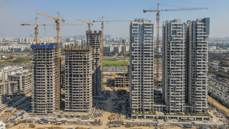 An aerial view of a sprawling construction site in an urban area, with multiple cranes and partially built high-rise residential towers alongside completed modern buildings.