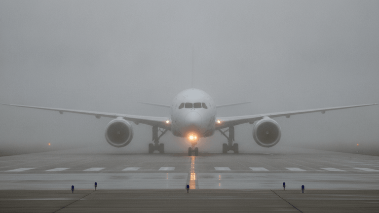 A large white commercial passenger jet sits on a runway, its front section visible through dense fog. Navigation lights are on.