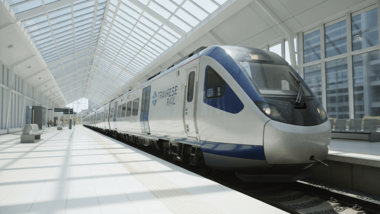 A modern silver and blue train with TRAVESE RAIL branding is parked at a clean, well-lit station platform.