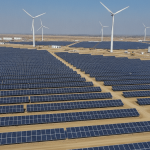 A wide shot of a vast renewable energy facility under clear daylight, featuring rows of solar panels, wind turbines, and battery storage.