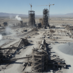 An aerial view of a vast industrial complex with multiple structures, cranes, and piles of material under clear daylight.