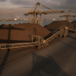 A wide, low-angle shot of large piles of iron ore at an industrial port with dormant cranes and conveyor belts.