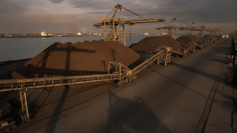 A wide, low-angle shot of large piles of iron ore at an industrial port with dormant cranes and conveyor belts.