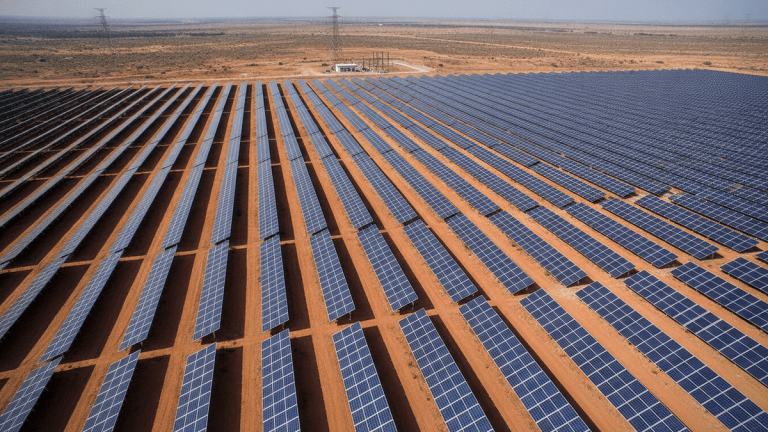 Aerial view of a vast solar power plant with rows of panels on reddish-brown earth, a substation, and transmission lines.