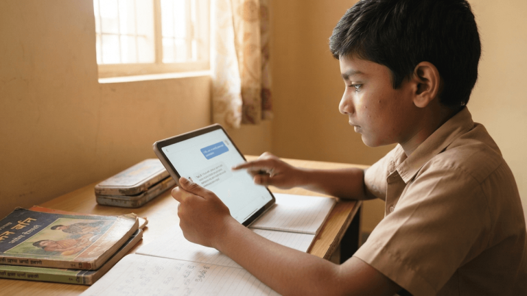 A 10-year-old Indian child touches a tablet screen displaying a generic AI tutor app, sitting at a wooden desk in a sunlit home.