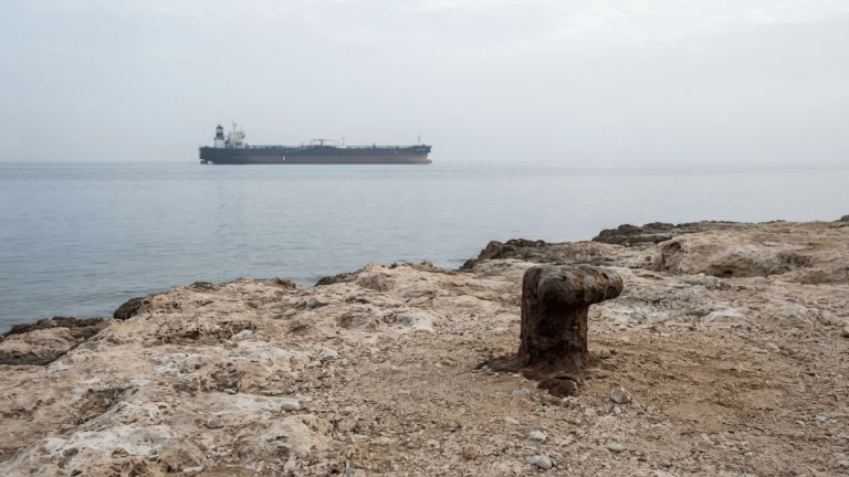 A rocky coastline of Perim Island with a large oil tanker visible on the horizon in the Bab el-Mandeb Strait.